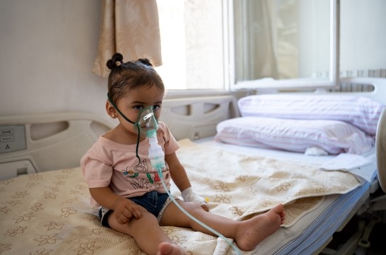 En liten jente sitter på en sykeseng og har på seg en pustemaske.