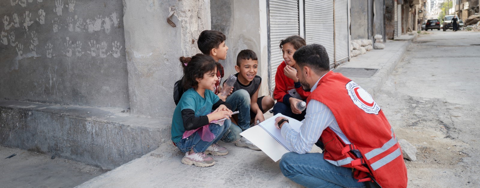 En frivillige fra Syrisk Røde Halvmåne snakker med barn som smiler og sitter på et fortau.