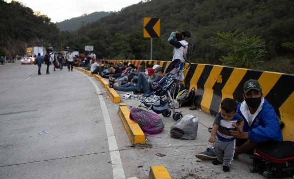 En gruppe migranter sitter langs en motorvei og tar en pause før de skal gå videre.