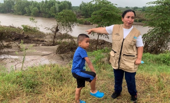 En kvinne står ute i gresset og peker på en elv bak seg. Ved siden av henne står det en liten gutt.
