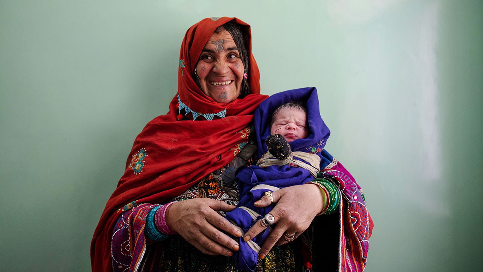 En Afghansk eldre Afghansk kvinne kledd i tradisjonelle fargerike klær med barnet i armkroken