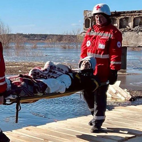 Røde Kors frivillig e bærer mann på båre, bro over vann, Ukraina