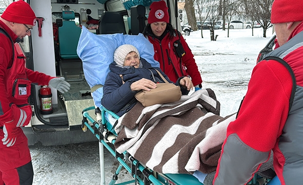En eldre kvinne på en båre hjelpes inn i en ambulanse av tre Røde Korsere.