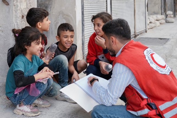 En mann fra Røde Halvmåne sitter på huk og snakker med barn som smiler.