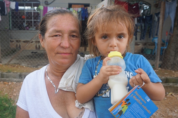 Et barn med tåteflaske blir holdt av en dame som ser i kamera.