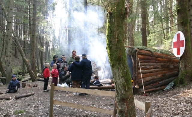 En gruppe barn står sammen noen voksne ute i en skog, ved en gapahuk. Røyk siver opp fra et bål som er tent.