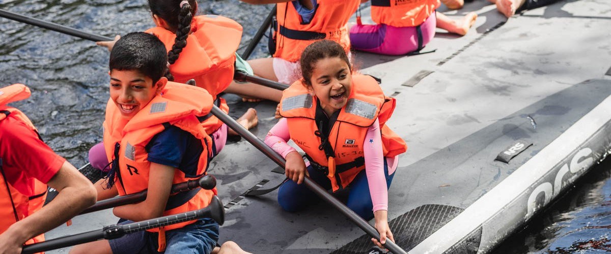 Mange barn sitter med redningsvester på et surfebrett og smiler