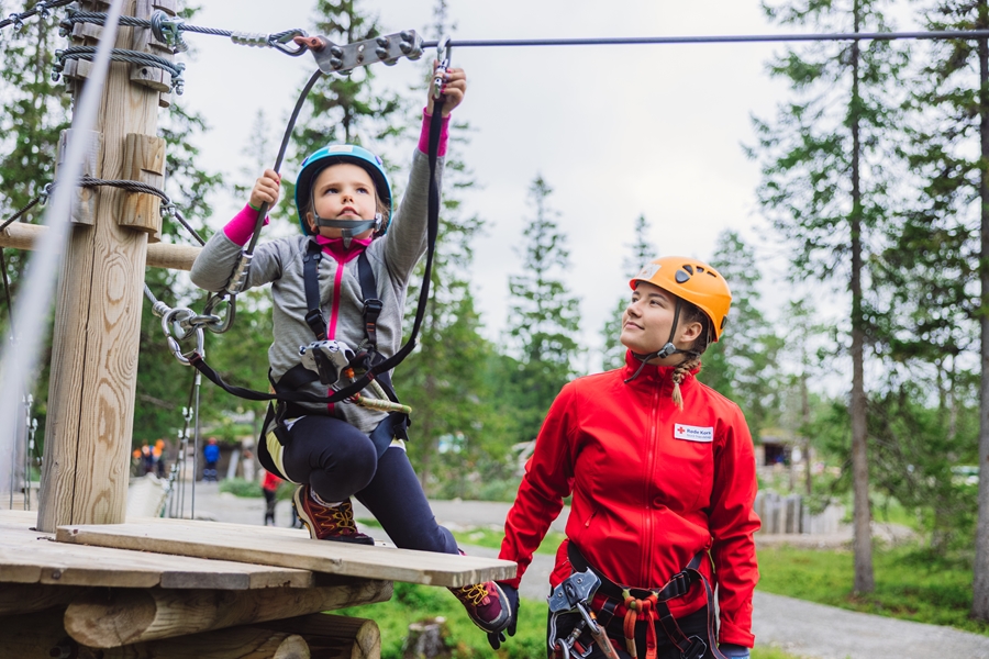 Jente fester sikring til vaier i klatreparkens, men en Røde Kors-frivillig følger med.