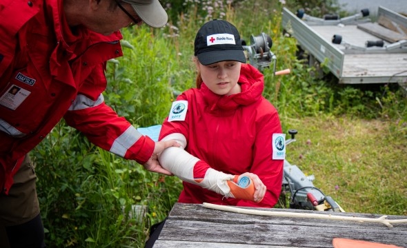 En jente sitter på bakken med en bandasje rundt armen. Hun øver på førstehjelp med bistand fra en voksen.
