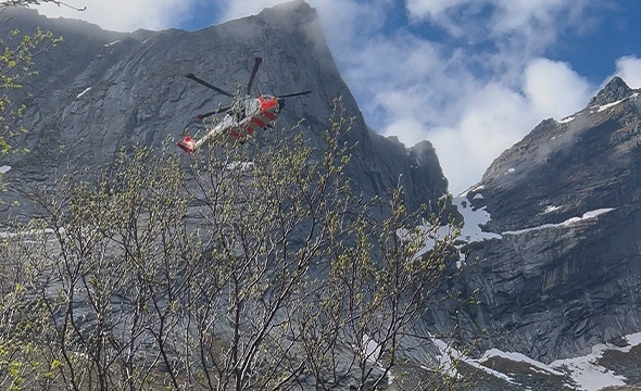 Et redningshelikopter sirkler i luften over området der de skal hente en skadet person. Det er fjell og trær i bakgrunnen.