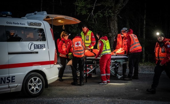 En ambulanse står klar og en gruppe frivillige løfter en båre inn med en skadet person.