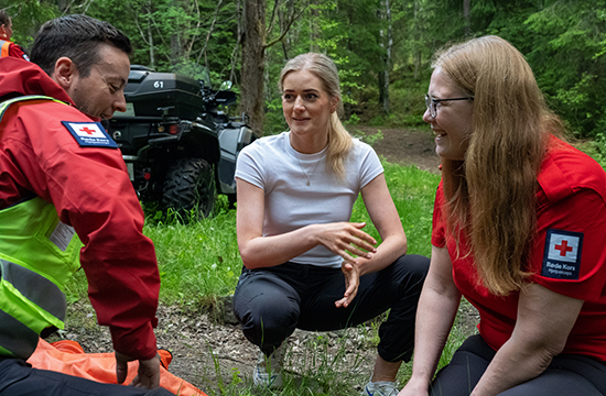 To personer fra Røde Kors sitter i en skog med en kvinne mellom seg