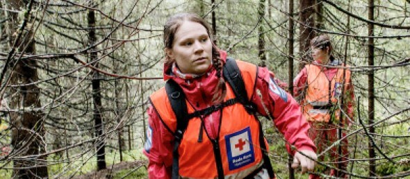 En ung kvinne i rød hjelpekorps uniform leter i tett gran skog