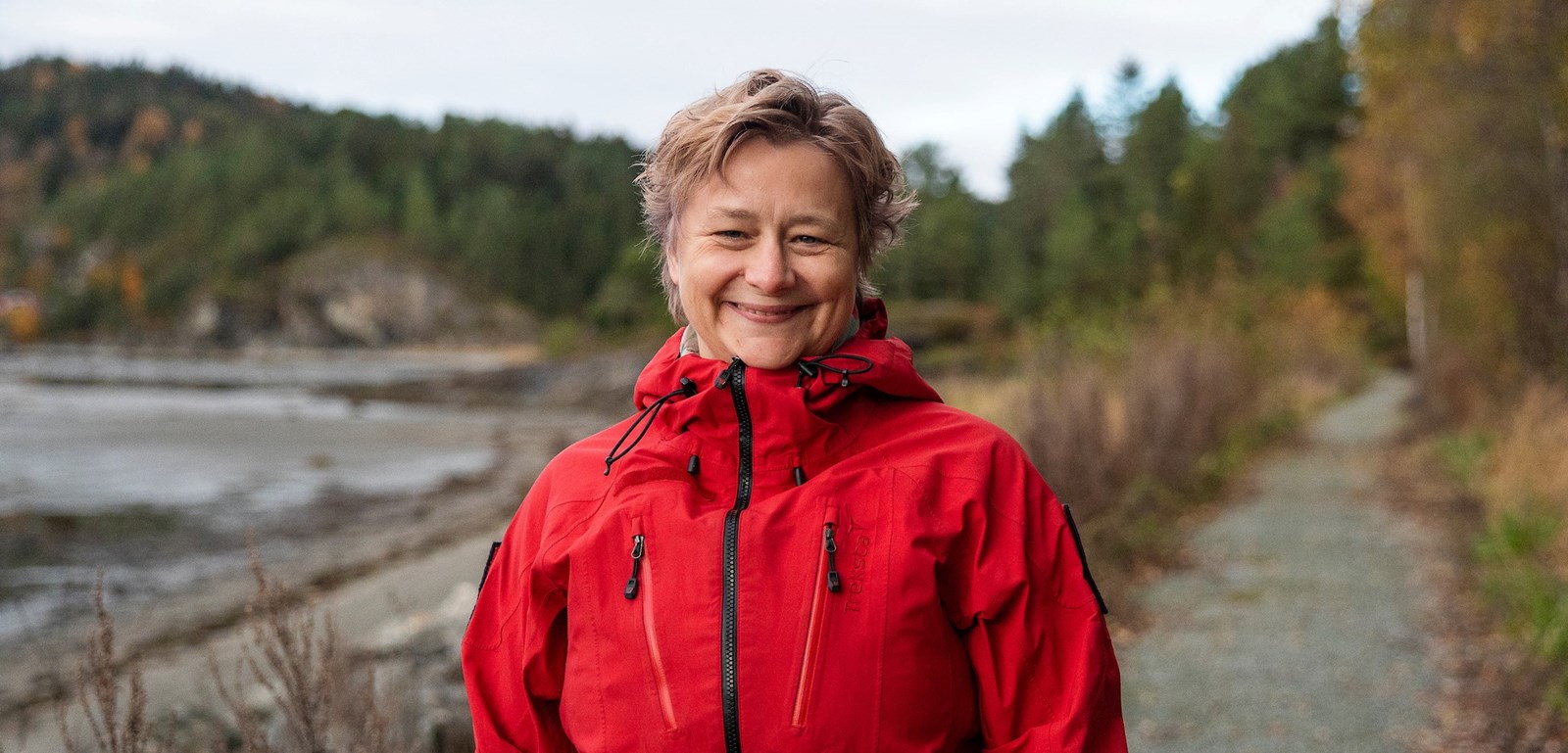 Portrettbilde av en smilende kvinne i rød jakke langs en skogsti