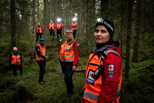 Kvinnelig hjelpekorpset med hodelykt ser i kameraet. Rundt henne er flere hjelpekorpsene i en grønn og mørk skog.