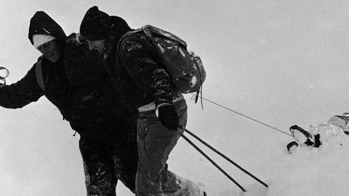 Svart hvitt-bilde av to personer som drar en tredje person, liggende, i tett snødrev på fjellet