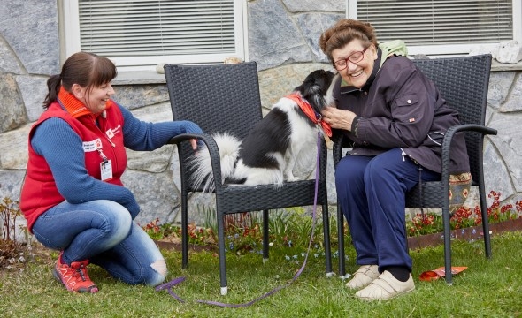 En eldre dame sitter på en stol med en hund ved siden av seg. Hunden slikker henne på kinnet og hun ler. På gresset ved siden av sitter en annen kvinne som eier hunden.