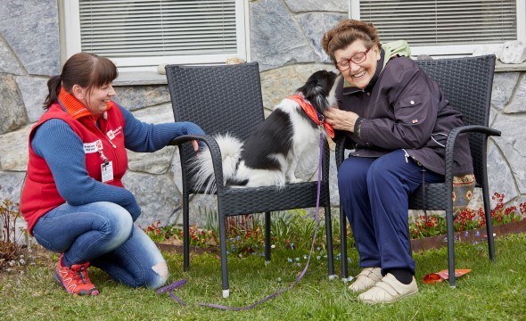 En eldre dame sitter på en stol med en hund ved siden av seg. Hunden slikker henne på kinnet og hun ler. På gresset ved siden av sitter en annen kvinne som eier hunden.