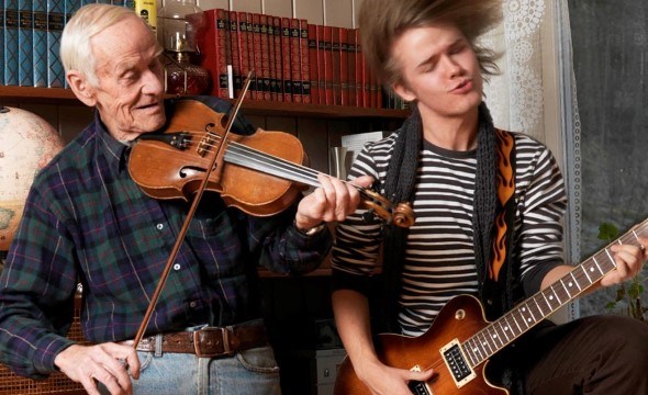 En eldre mann spiller fele i en sofa sammen en yngre mann som spiller på elektrisk gitar. De er fulle av liv og lever seg inn i musikken.