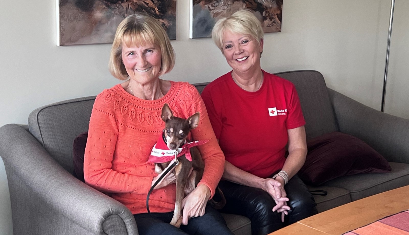 En kvinnelig besøksvenn sitter med hunden sin på fanget, og smiler til kameraet sammen med sin besøksvert.
