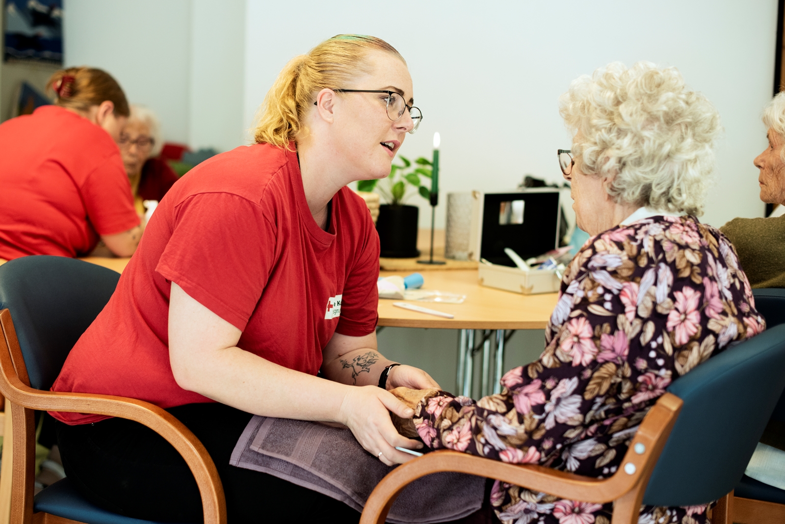 To personer sitter i rom på et sykehjem i en dyp samtale. Den yngre damen har Røde Kors-klær og holder den eldre damen i hendene.