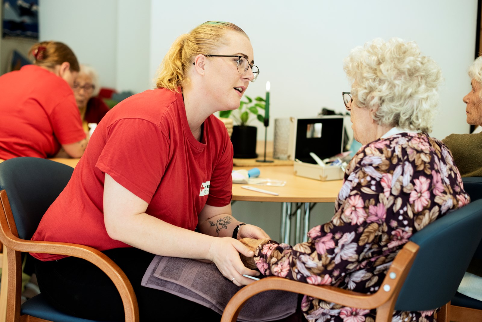 To personer sitter i rom på et sykehjem i en dyp samtale. Den yngre damen har Røde Kors-klær og holder den eldre damen i hendene.