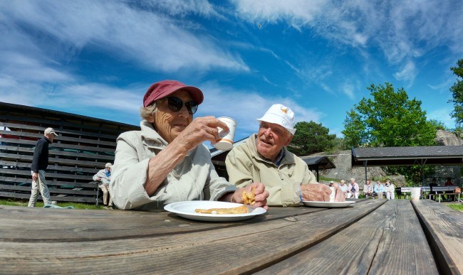 En eldre mann og en eldre kvinne sitter ute og spiser vaffel og drikker kaffe