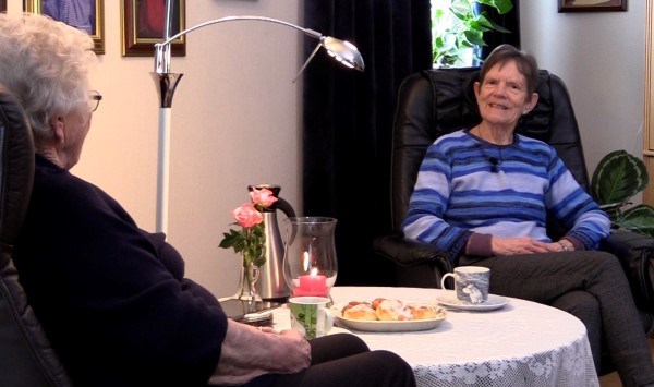 To eldre hvinner sitter i hver sin stol mens de drikker kaffe. Det står wienerbrød på bordet.