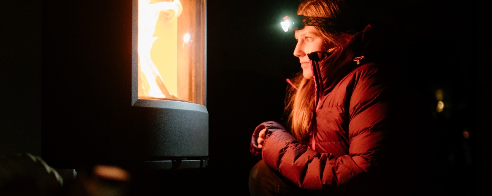 En kvinne med boblejakke og hodelykt sitter foran en vedovn der ilden flammer. 