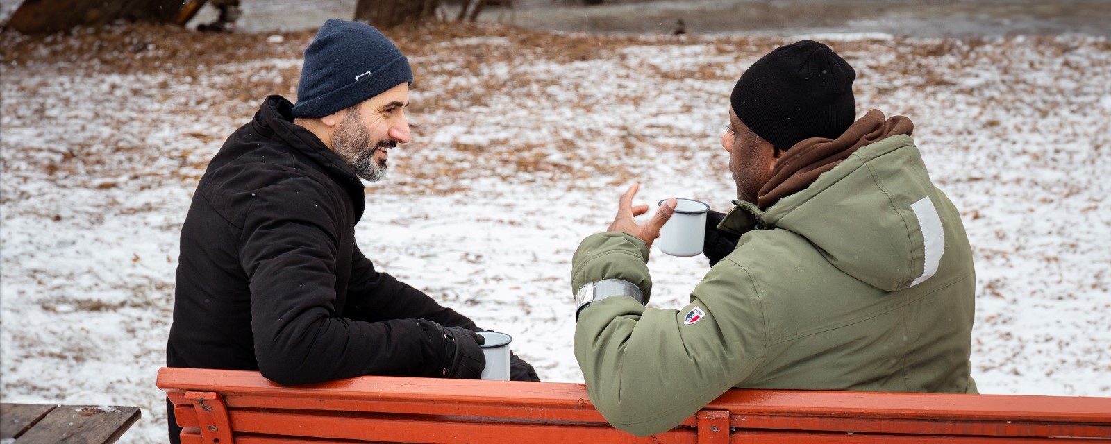 To menn sitter på en benk ute i en park på vinterstid . De snakker sammen og drikker noe  fra to kopper.